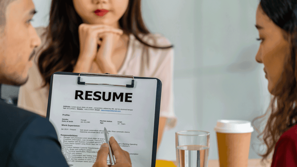 A resume on a clipboard on the table between three people in a meeting