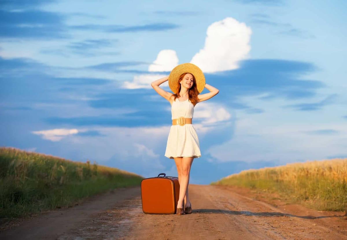 A woman in a sunhat standing on a country road beside a suitcase