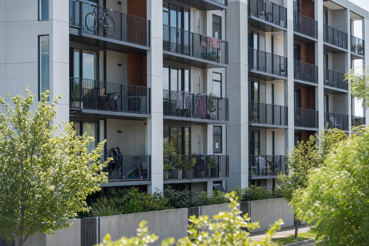 A low-rise purpose-built rental apartment building in a New Zealand city on a bright morning, bicycles and planters on the balconies