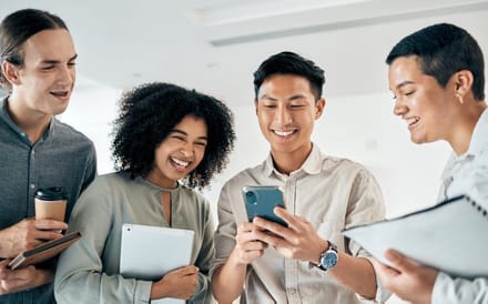 Four colleagues standing together, looking at a phone and holding tablets and coffee
