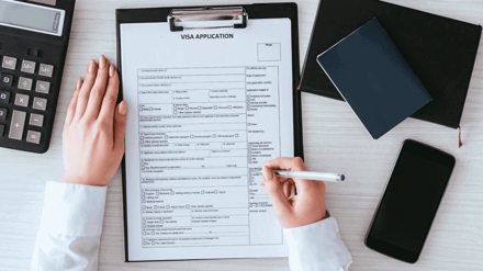 Hands filling in a visa application form on a clipboard, phone and notebook alongside