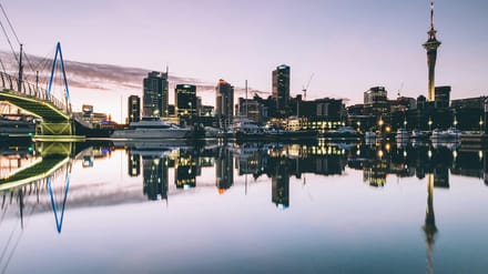 Auckland harbour at dusk, city towers and construction cranes reflected in still water, an ordinary working skyline.