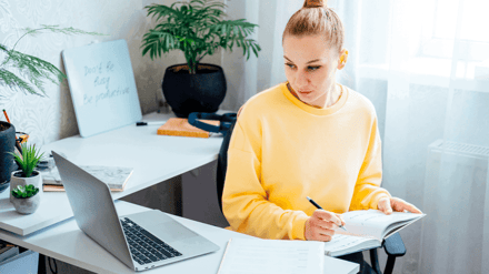 A woman in a yellow jumper taking notes at a desk beside a laptop