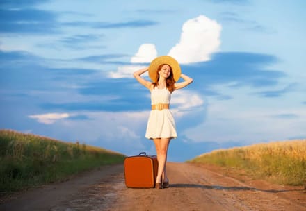 A woman in a sunhat standing on a country road beside a suitcase