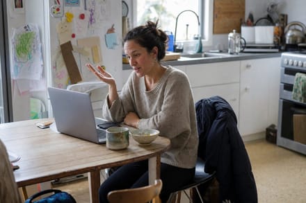 Two women talking across a kitchen table with mugs and an open notebook between them.