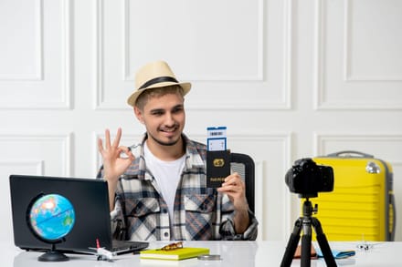 A man at a desk with a passport, suitcase and camera, giving an OK sign