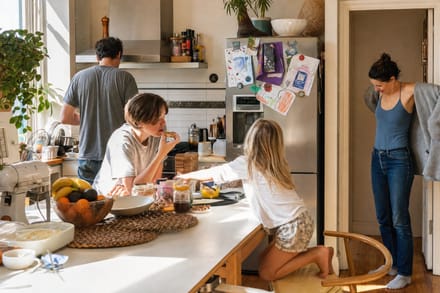 A family of four walking away along a suburban footpath, backpacks on, weatherboard houses either side.