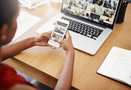 Hands holding a phone beside a laptop showing a video call
