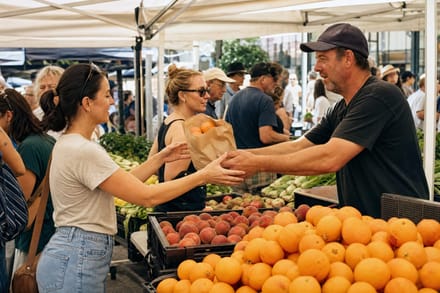 A busy weekend farmers market, people browsing produce stalls under white canopies.