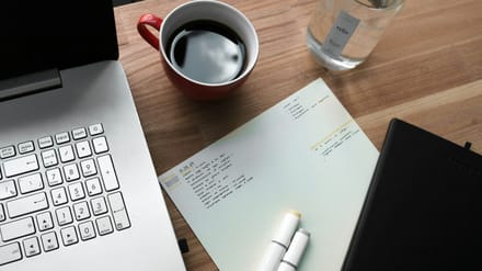 A desk with a keyboard, a notepad, a pen and a cup of coffee